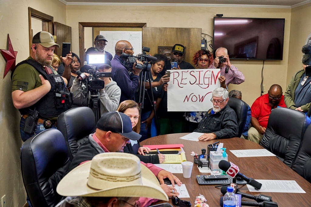 A person holds a sign reading "Resign Now" inside a meeting with press cameras. 
