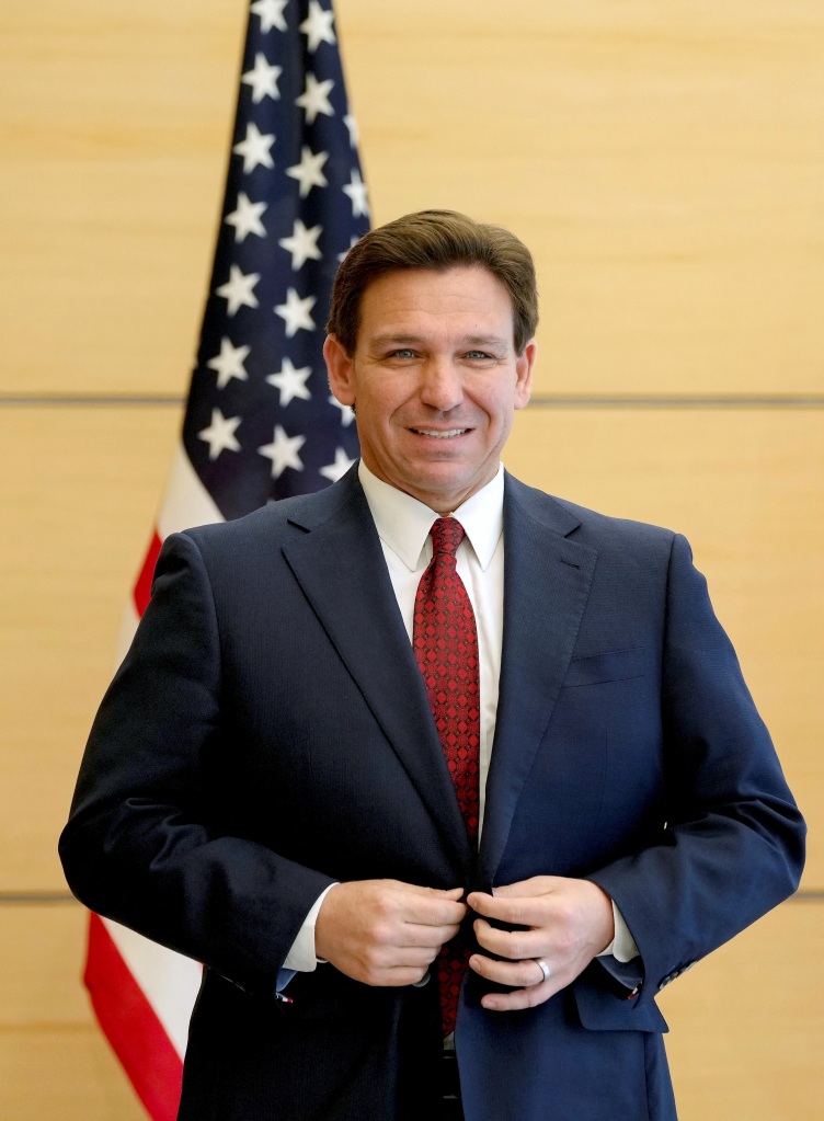 Gov. Ron DeSantis buttons his suit jacket as he stands in front of an American flag. 