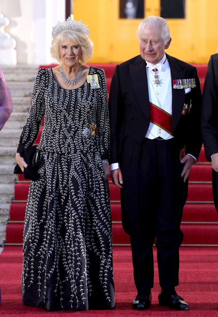 King Charles III and Camilla, Queen Consort, smile standing together on a red carpet. 