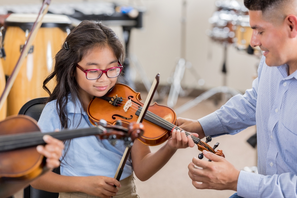 A smiling little girl sits next to her attentive music teacher and plays the violin during music class.  