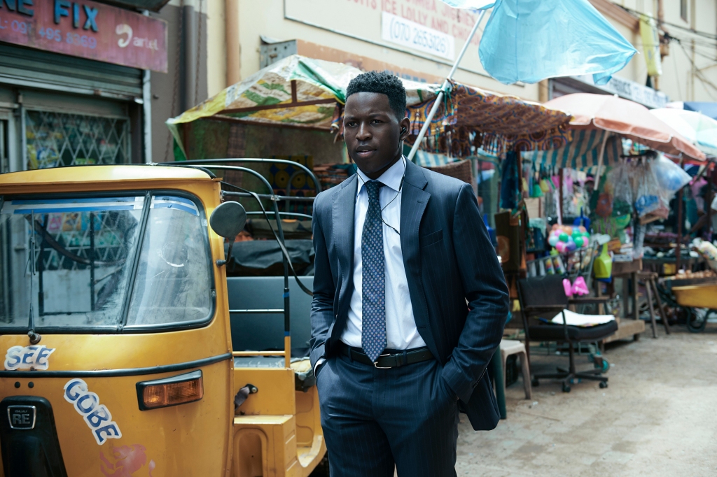 Toheeb Jimoh as Tunde. He's standing on a street in Lagos and is wearing a business suit and headphones. His hands are in his pockets.