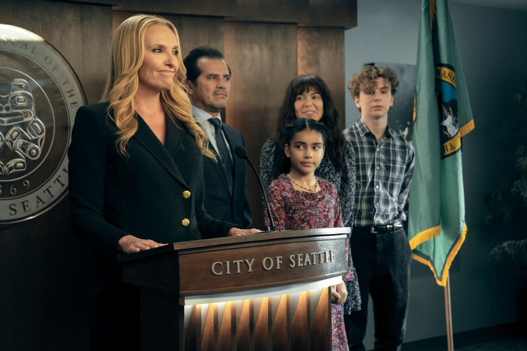 Toni Collette as Seattle mayor Margot Cleary-Lopez, who's also Jos' mother. She's standing at a lectern and smiling during a press conference, with her family by her side.