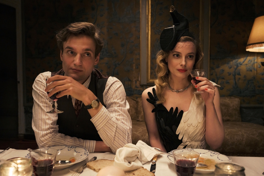 Lucas Englander and Gillian Jacobs as Albert and Mary Jayne. They're seated at a dinner table and are both dressed nicely. She's wearing a dress that looks like it's made from gloves and a black hat. They're both holding wine glasses and looking off-camera.
