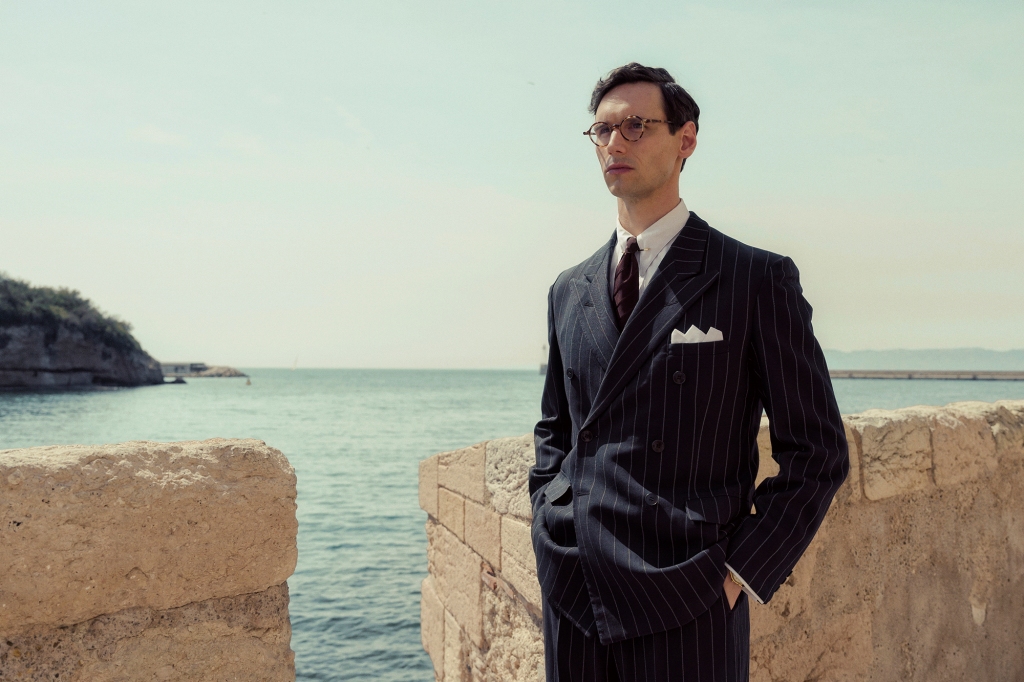 Cory Michael Smith as Varian Fry. He's wearing a pin-striped suit and glasses and is standing in front of an ocean-front stone wall with his hands in his pockets.