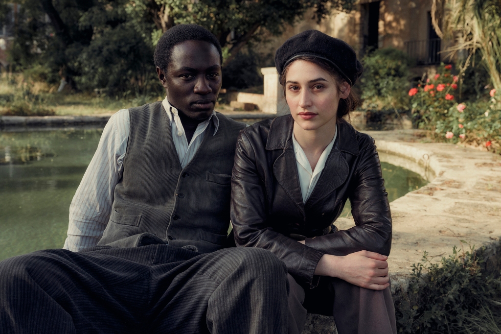 Ralph Amoussou and Deleila Piasko as Paul and Lisa in "Transatlantic." They're sitting next to each other on a stone wall and are looking directly into the camera. He's wearing a grey vest and a long-sleeve shirt; she's wearing a leather jacket and a black beret with her arm on her knee.