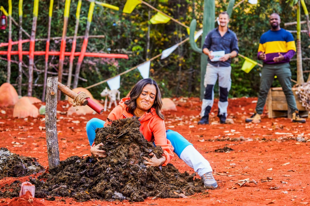 A young woman is outside with her arms around around a pile of black-and-brown goo that she looks like she's trying to pick up. She's grimacing and her eyes are closed. Bear Grylls and Jordan Conley are in the background, watching.