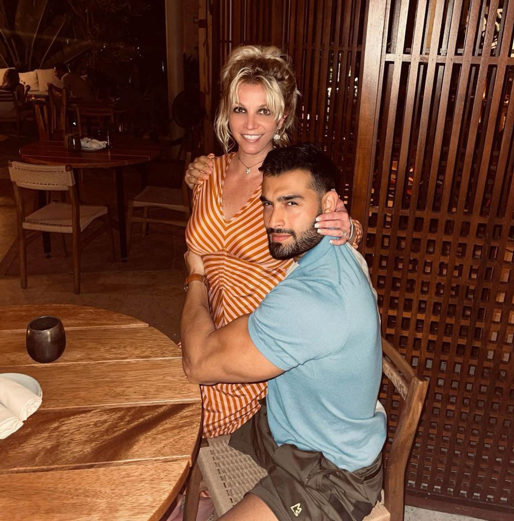 Britney Spears stands while hugging Sam Asghari, who is sitting. 