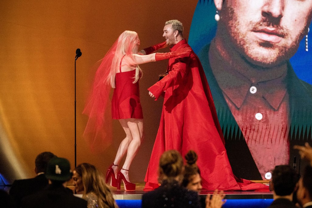 Blonde woman in red mini dress and man in red cape with long sleeves on stage at the 2023 Grammy Awards.