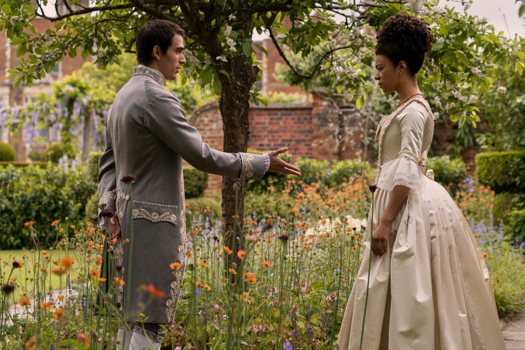 Corey Mylchreest as Young King George, India Amarteifio as Young Queen Charlotte, in "Queen Charlotte." They stand  in a garden, facing each other. 
