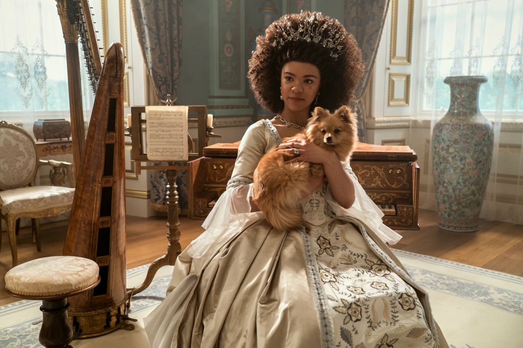 The younger version of Queen Charlotte, played by India Amarteifio, wearing a gown holding a dog in a fancy sitting room. 