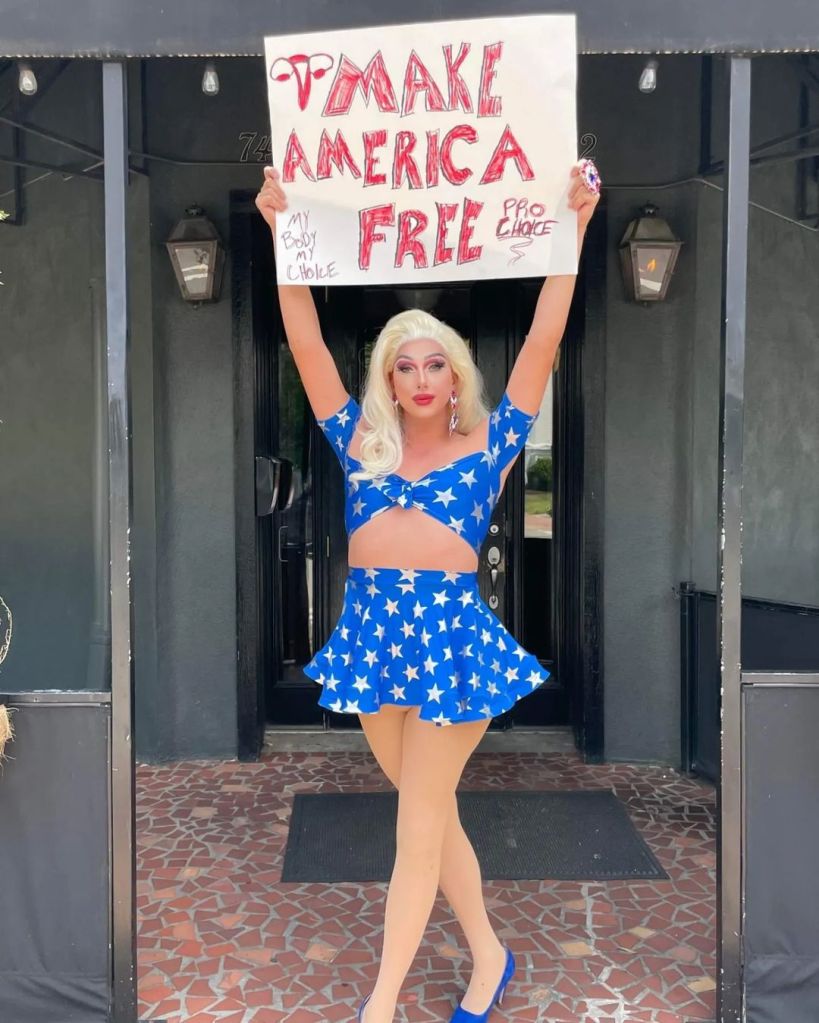 Joshua Kelley holding a sign reading "Make America Free" while dressed in drag in a matching skirt and crop top set.