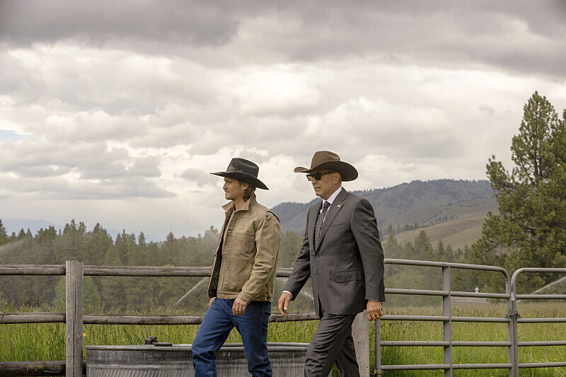 Father and son: Kayce (Luke Grimes) and dad John Dutton (Kevin Costner) in "Yellowstone" walk next to a field wearing cowboy hats. 