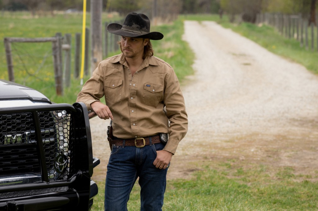 Luke Grimes in a cowboy hat leaning on an SUV. 