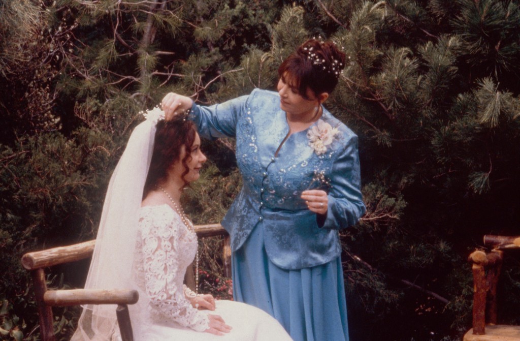 Sara Gilbert sitting on a bench in a bride outfit with a veil, while Roseanne Barr stands above her. 