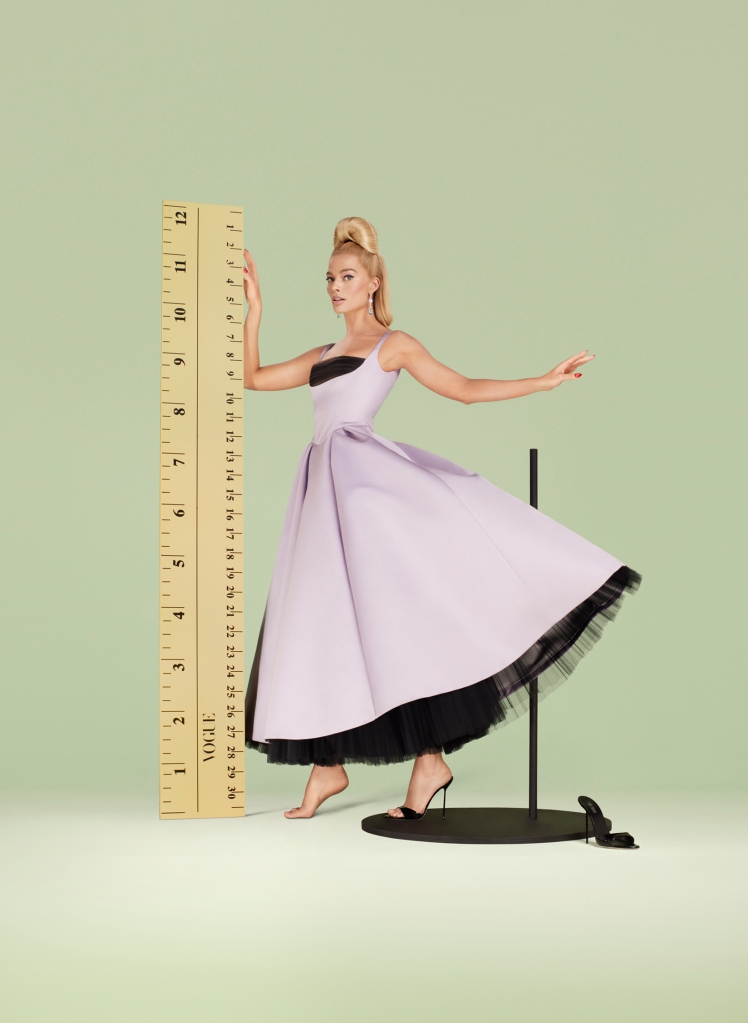 Margot Robbie in purple pastel gown next to a life-sized ruler on green back drop