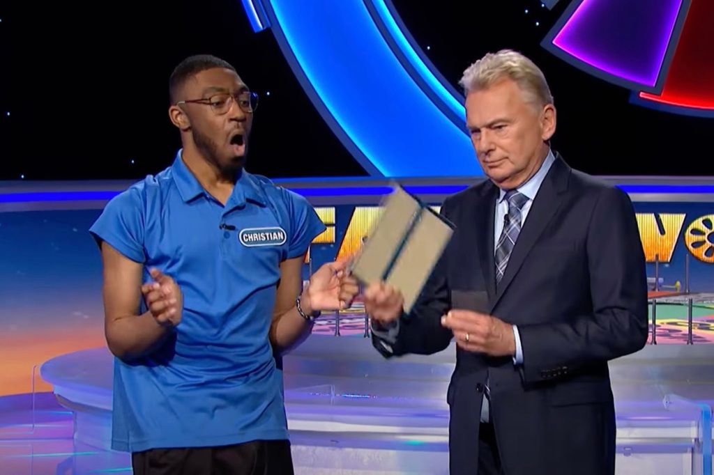 Pat Sajak and contestant Christian Dixie, who's trying to grab the envelope out of Pat's hand as Pat looks grumpy.