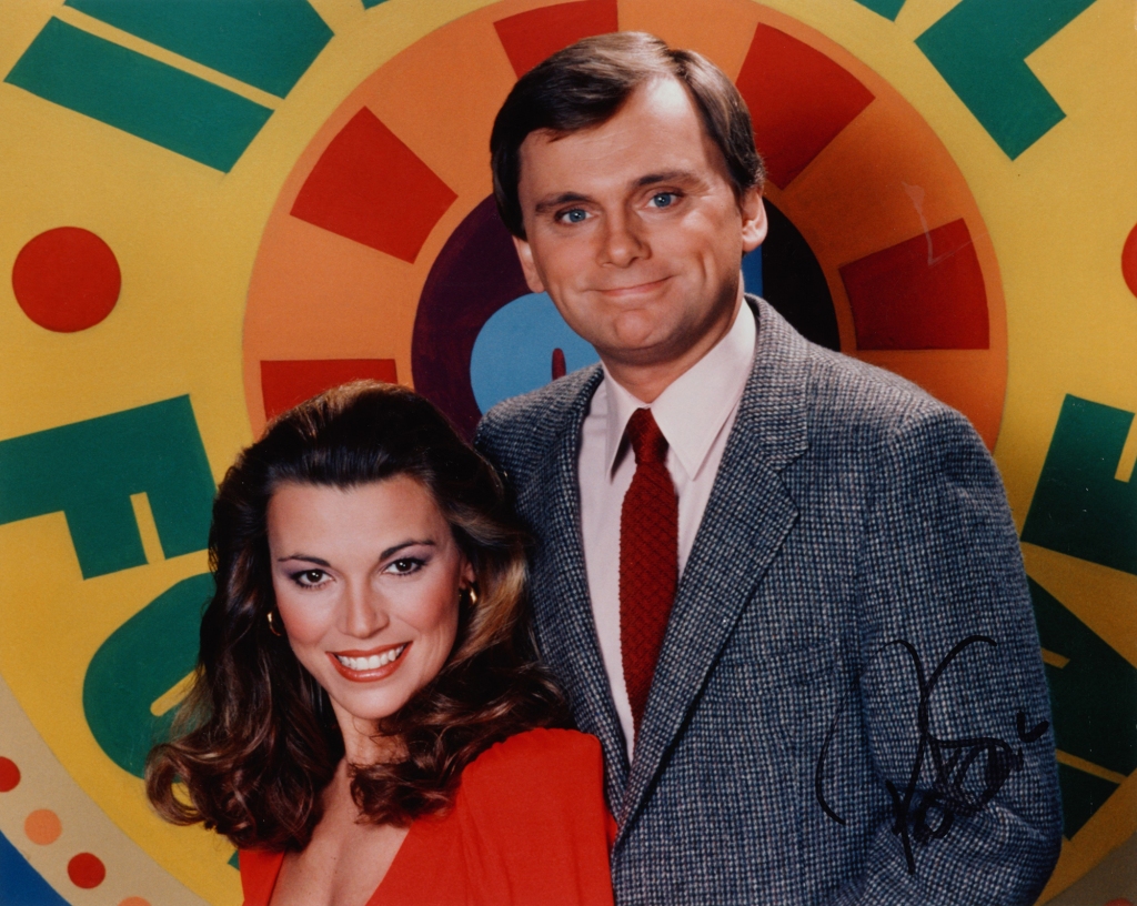 Photo of Vanna White and Pat Sajak from 1975. Vanna is wearing a red dress and has dark hair; Pat is wearing a blue checked sports jacket with a red tie and also had dark hair.