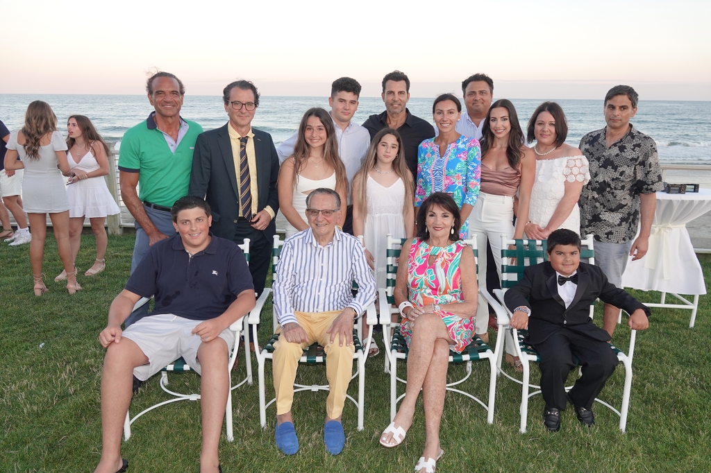 Frank and Joyce Caprio pose with their large family. They're sitting on lawn chairs on the grass with the ocean behind them.