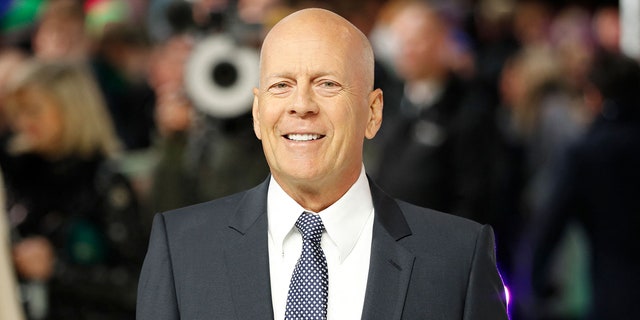 Bruce Willis smiles on the red carpet wearing a black suit, white button down and blue patterned tie