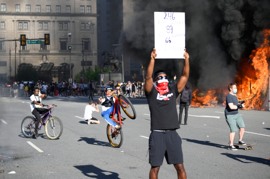 A man poses with a sign in front of burning police vehicles while protestors clash during the George Floyd riots in Philadelphia on May 30, 2020. 