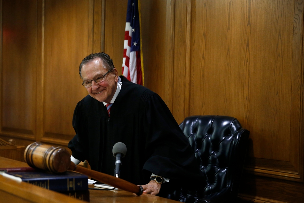 Photo of Judge Frank Caprio in his courtroom. He's standing up and leaning forward and smiling. There's a gavel in the foreground. He's wearing his black judge's robe.