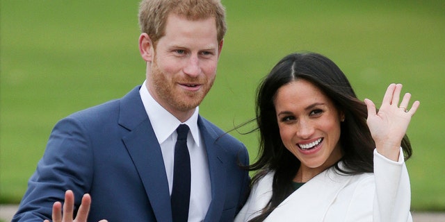 Prince Harry in a blue suit waves for photos as he walks out with his new fiancée Meghan Markle, wearing white and putting her hand up in the air