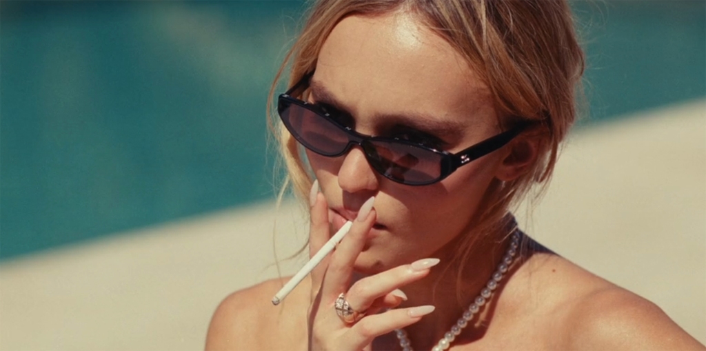 Lily Rose Depp wearing sunglasses, smoking by a pool. 