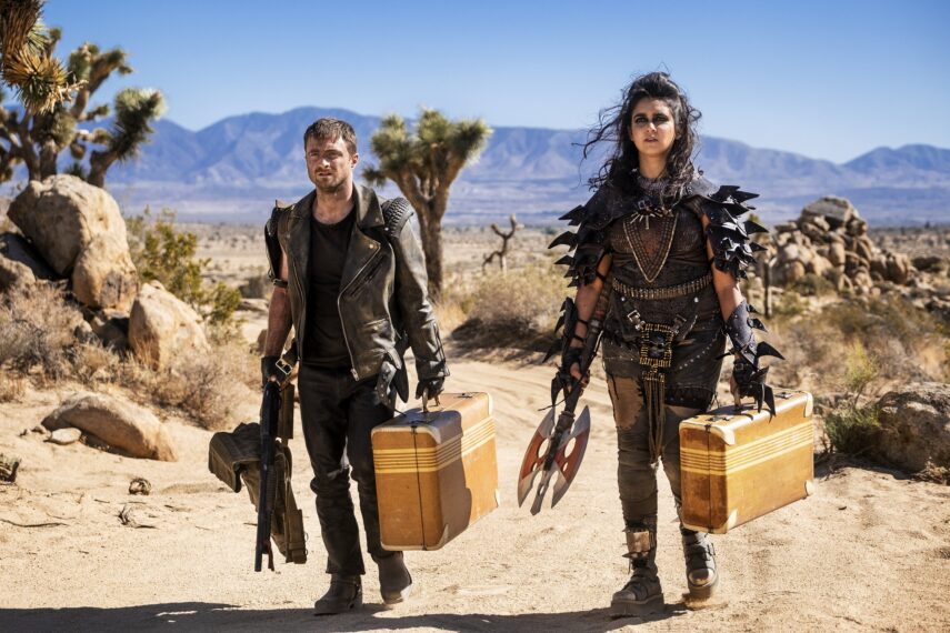 Daniel Radcliffe and Geraldine Viswanathan holding suitcases wearing leather standing in a desert. 