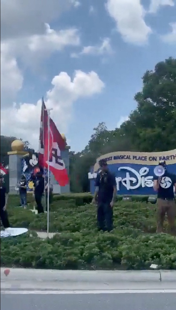 People stand next to large red Nazi flags and a man speaks into a megaphone at the entrance to Walt Disney World. 