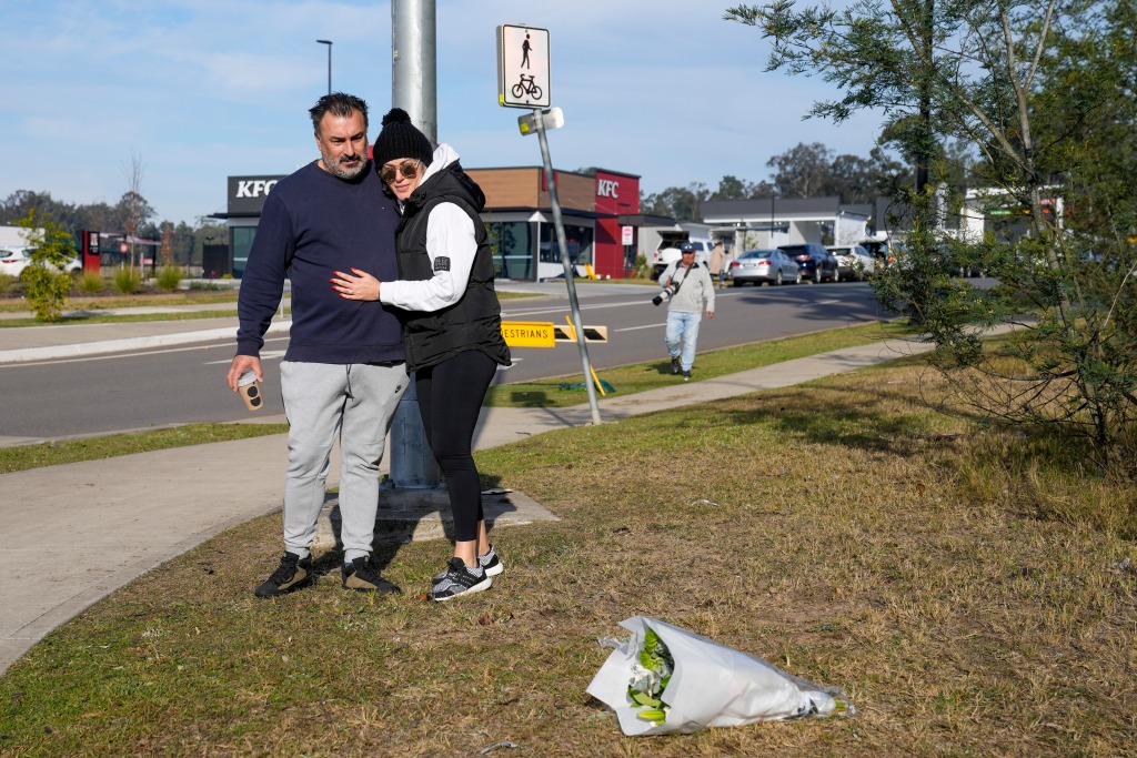 A woman hugs a man as they look at a bouquet of flowers place on a grassy area on the side of a road. 