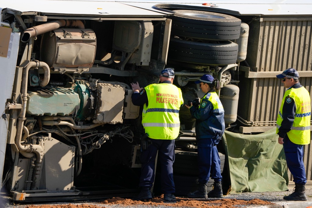 Police inspect the underside of a flipped bus lying on its side. 