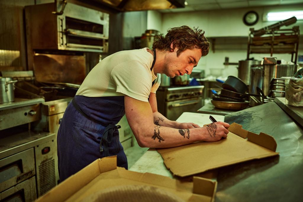 Jeremy Allen White in a kitchen bent over, writing. 