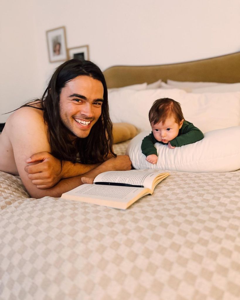 Man with long dark hair lying on bed smiling at camera next to baby.