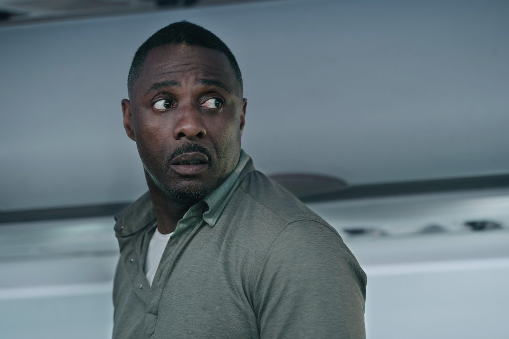 Idris Elba standing on a plane looking worried. 