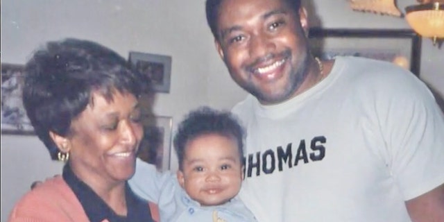 Minnie Smith is holding a smiling baby next to her smiling grandson wearing a grey sweatshirt