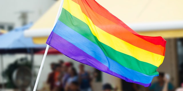 Rainbow flag waving on the street during a gay pride celebration with unrecognizable people lining the sidewalk in the background. U.S. embassies can now display the flag on the same pole as the American flag in June, during Pride month.