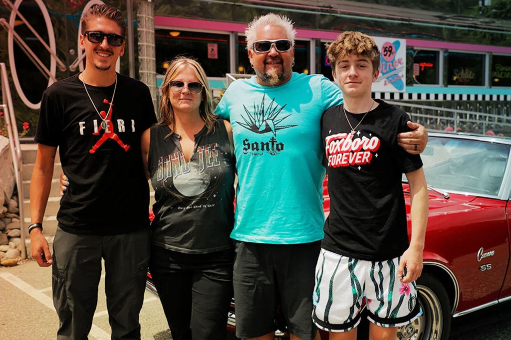 Guy Fieri poses with his wife Lori and their sons Hunter and Ryder. They're standing side-by-side outside in front of a vintage red Camaro. Guy's got his arms around his wife and son.