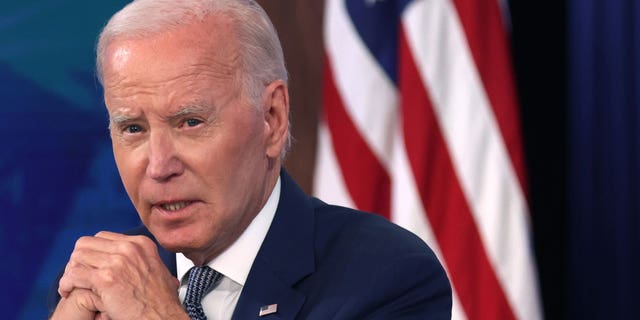 President Joe Biden holds his hands together as he sits in front of an American flag