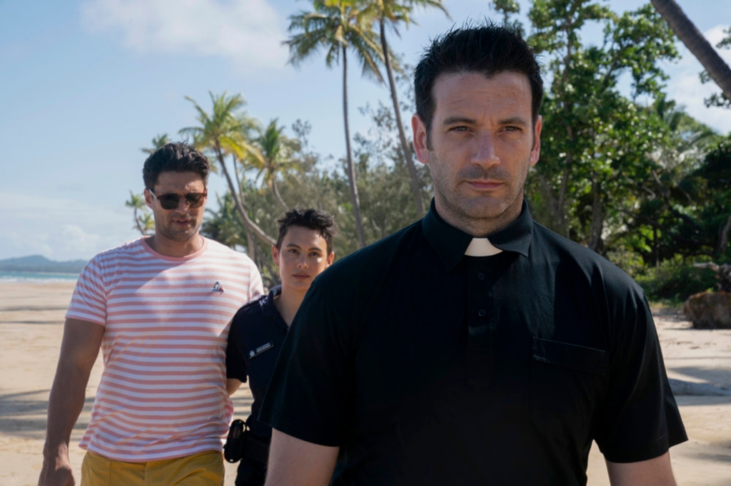 Colin Donnell as a fake reverend in "Irreverent." He's standing on a sandy beach with palm trees and wearing a black short-sleeved shirt and a white clerical collar.