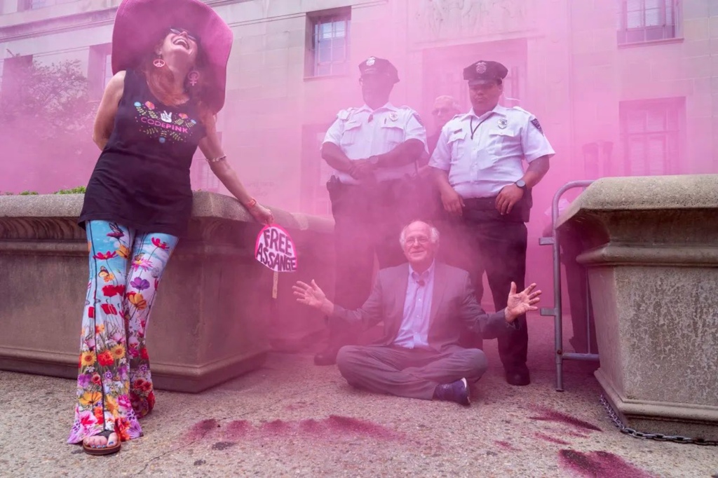 Ben Cohen sits in the pathway to a DOJ building in front of two officers as pink smoke fills the air. 