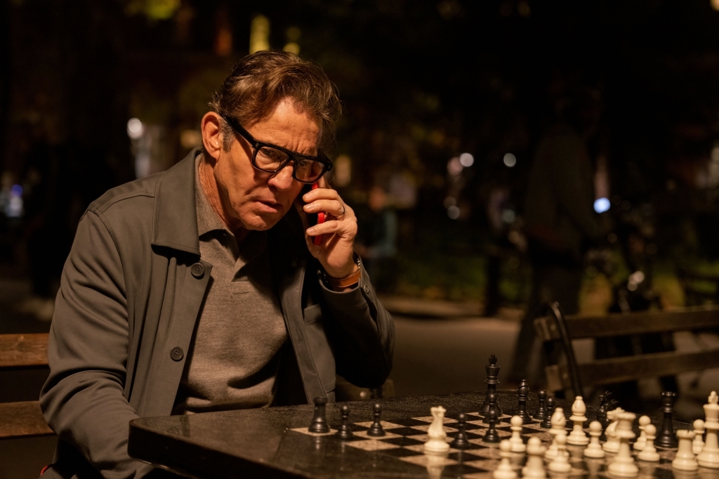 Dennis Quaid on a phone at a chessboard outside. 