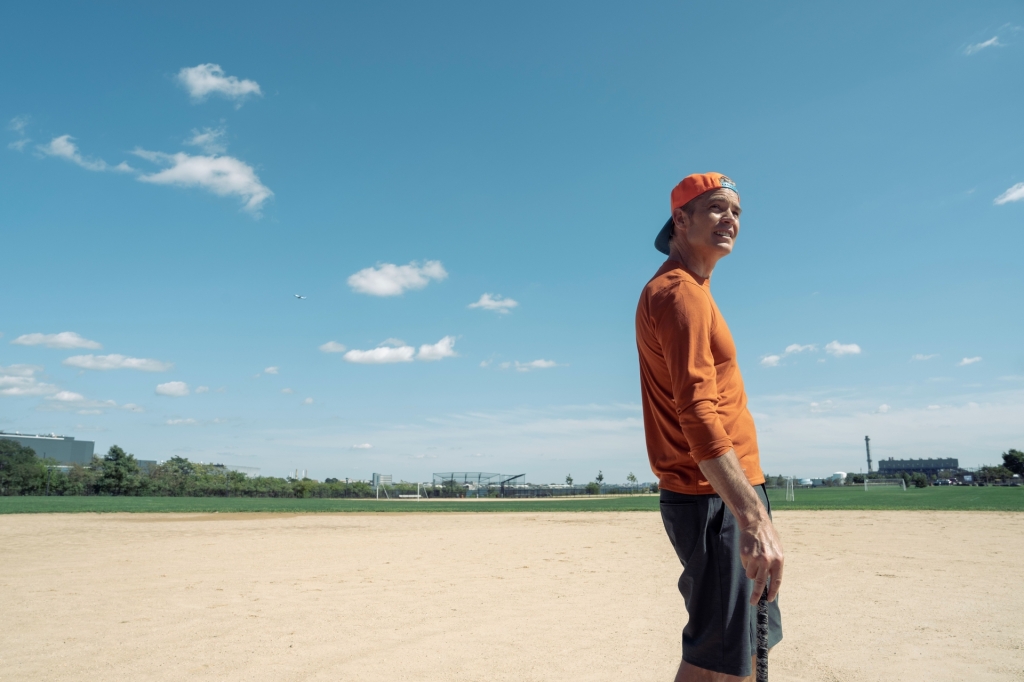 Timothy Olyphant on a baseball field in a backwards baseball cap. 