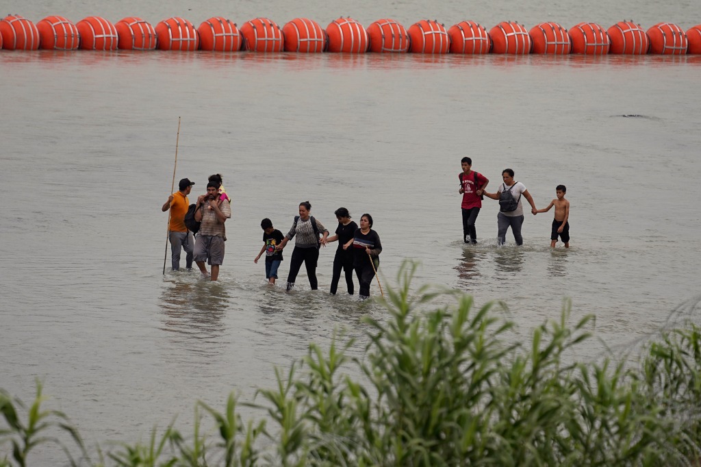 Migrants who crossed the Rio Grande from Mexico walk past large buoys being deployed as a border barrier