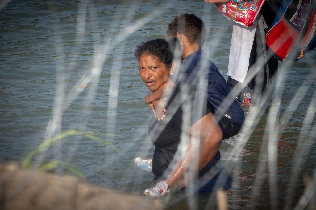 A woman holds a child on her back as migrants walk near concertina wire in the water along the Rio Grande border