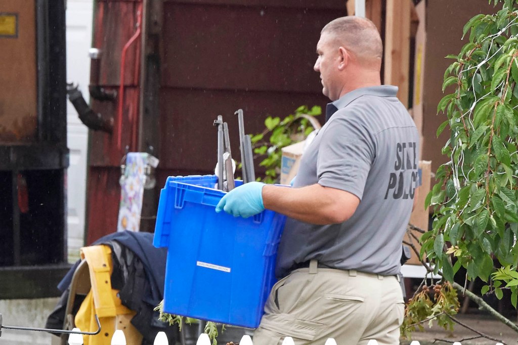 State police officer carries a blue bin of items out past house.