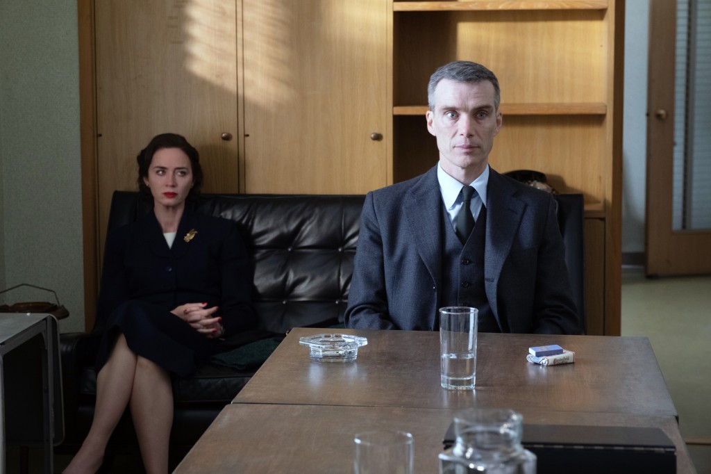 Emily Blunt on a couch while Cillian Murphy sits at a table near her. Both look serious. 