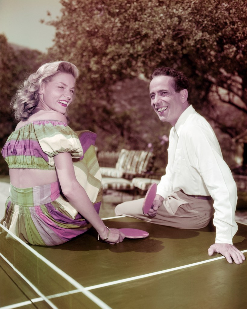 Humphrey Bogart and Lauren Bacall sitting on ping pong table with paddles.