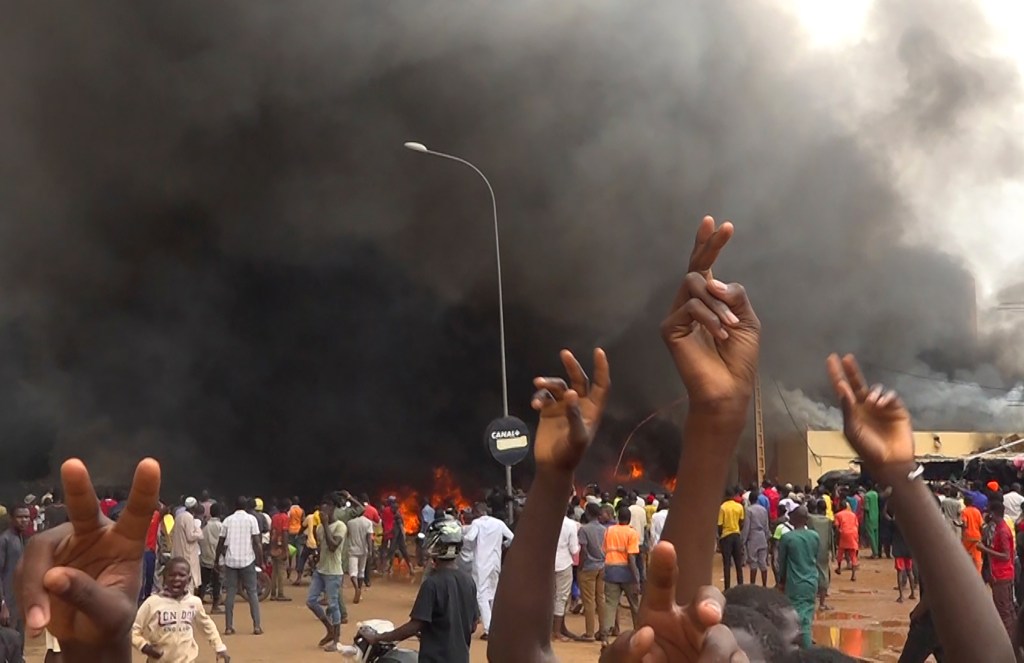 The headquarters of Niger's ruling party burns in the background as supporters mo mutinous soldiers demonstrate in Niamey, Niger.