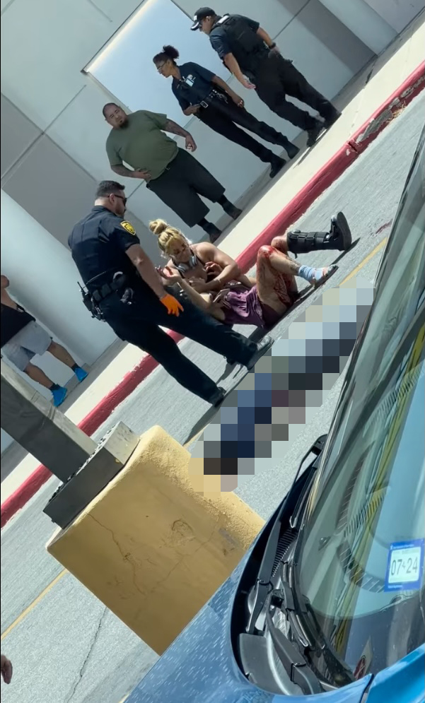 In photos shot at the scene, the female thief is seen being assisted by a police officer and another female with blood running down her leg, while the dead thief is laying face down in the parking lot. 