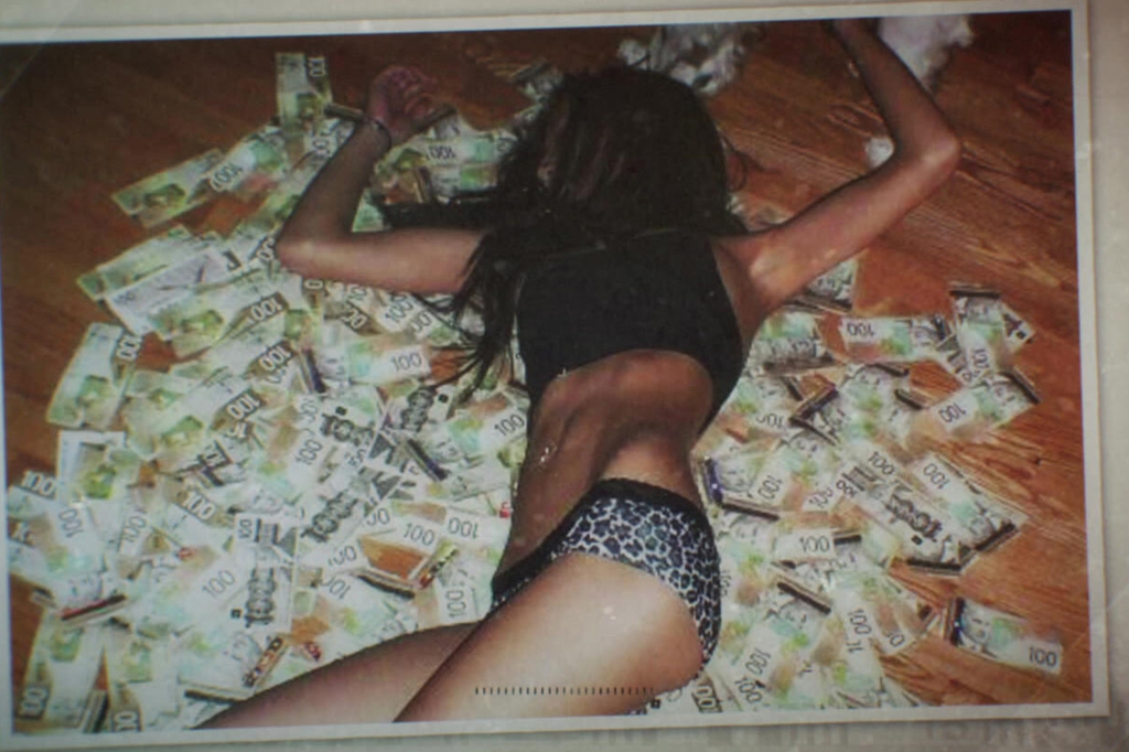 Polaroid-type photo of a scantily clad woman lying on her back on a mountain of 100-dollar bills. She's wearing a black top and leopard-skin underwear. Her face is hidden by her hair.